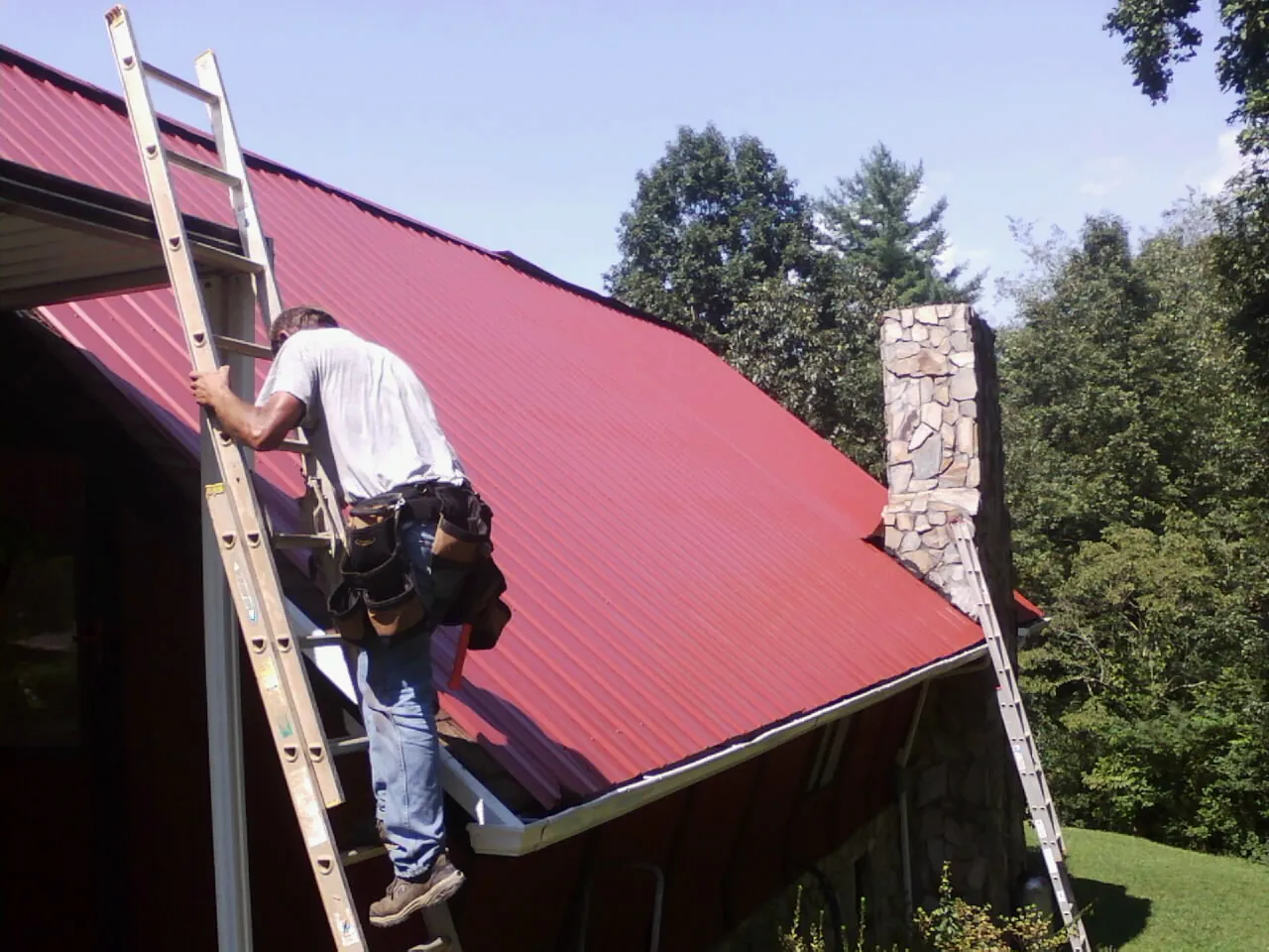 Roofer on a ladder beside a red metal roof and stone chimney — Soffit & Fascia Repair by Roofing Brothers in Cumming